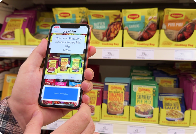 A smartphone held up in front of supermarket shelves stocked with Colman’s meal mix packets. The phone screen uses Zapvision technology to detect and label “Colman’s Singapore Noodles Recipe Mix 24g” located 56 cm away.