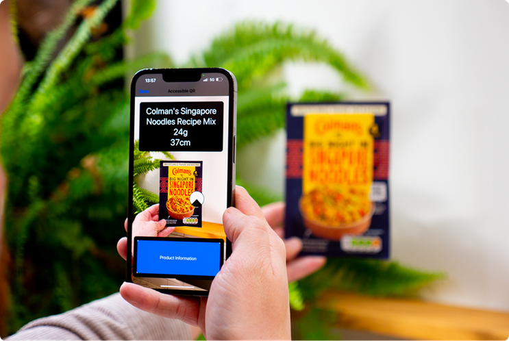 A hand holding a smartphone in front of a Colman’s Singapore Noodles Recipe Mix packet, with green ferns in the background. The phone uses Zapvision technology to identify the product as “Colman’s Singapore Noodles Recipe Mix 24g” located 37 cm away, displaying the result on-screen along with a “Product Information” button.