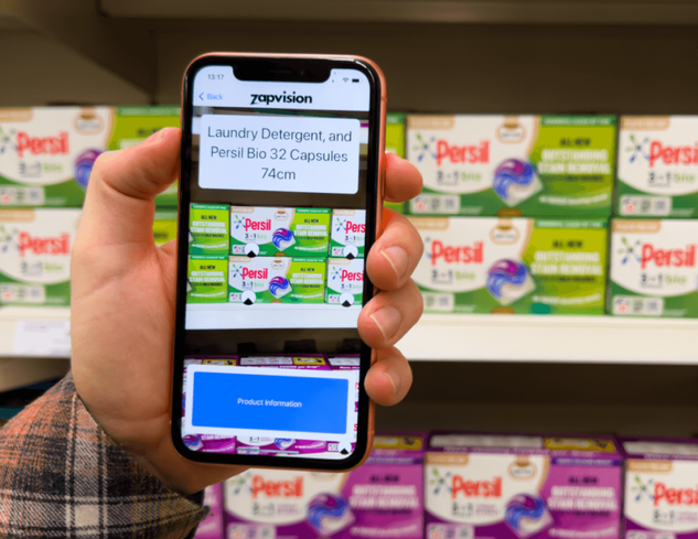 Person holding a mobile device showing the Zapvision app, scanning persil bio 32 capsules packaging.